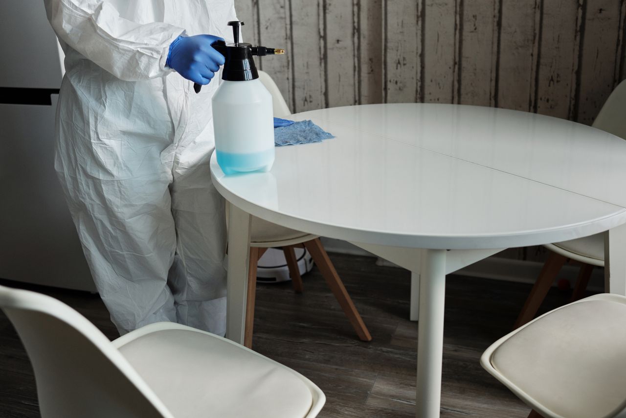 services-02 Person in protective suit disinfecting a white table indoors with a sprayer.